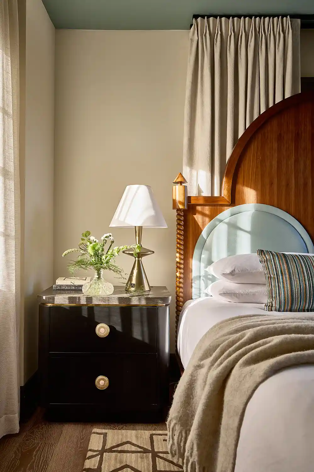 Styled bedroom with a made bed, wooden headboard and bedside table with lamp and flowers.